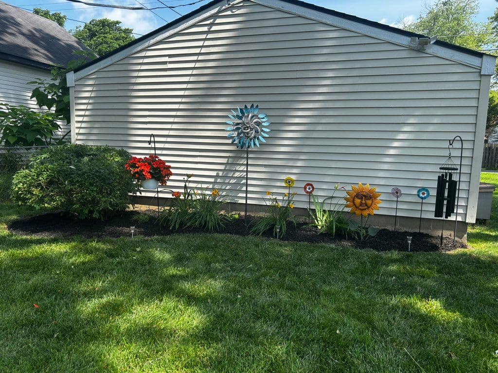 Mulched flower bed along garage wall with plants and decorative garden stakes